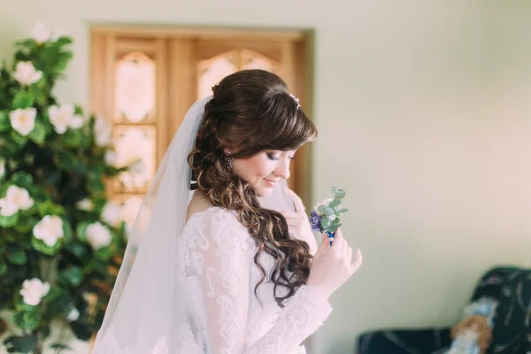 Beautiful bride with long curly hair and veil looking at the buttonhole indoors — Stock Photo, Image