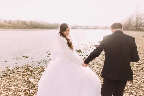 Mladý pár se právě oženil a drží se za ruce na břehu řeky — Stock fotografie