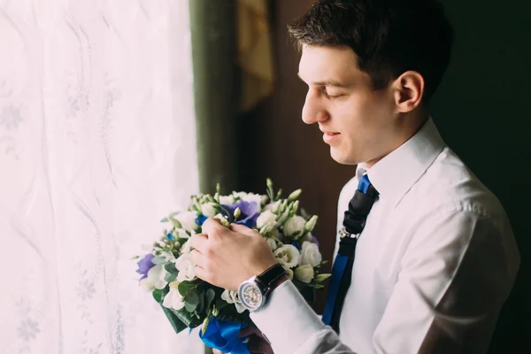 Elegante bonito noivo de cabelos escuros segurando um buquê de casamento em pé perto da janela — Fotografia de Stock