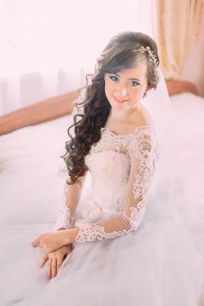 Young beautiful bride with long curly hair sitting in his bedroom at edge of the bed and smiling — ストック写真