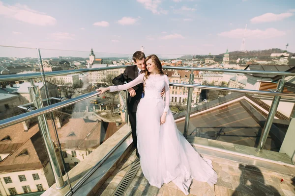 Красивый жених и невеста обнимаются на крыше здания с городским фоном — стоковое фото
