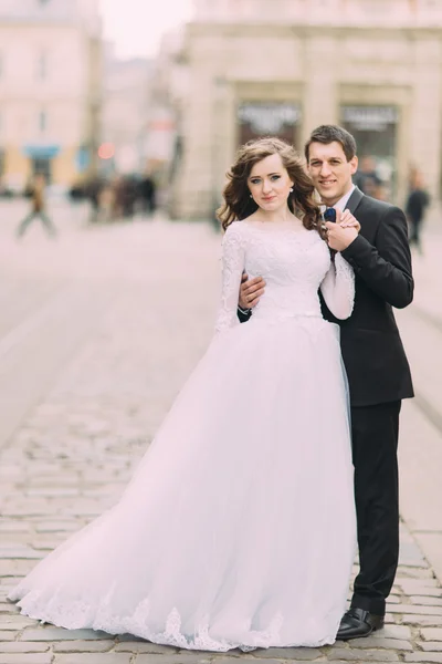 Bonito noivo em terno preto segura a mão da noiva vestindo vestido branco, velho fundo ensolarado da cidade — Fotografia de Stock