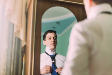 Otel odasında aynaya karşı dururken kravatını ayarlayan ve gülümseyen formalgiyimli yakışıklı genç adam