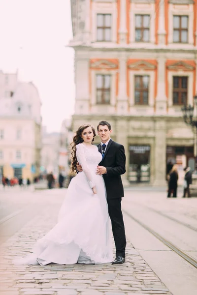 Boldog menyasszony és a vőlegény stílusos futás ölelgetés a háttérben régi napsütötte város — Stock Fotó