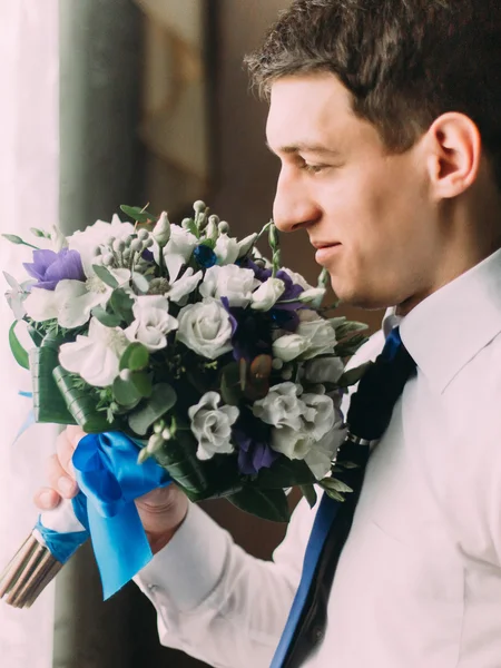 Elegante bonito noivo de cabelos escuros cheirando um buquê de casamento em pé perto da janela — Fotografia de Stock