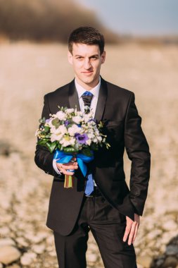 Şık şık gülümseyen genç damat buket çiçek yalnız rocky Sahilde yürüyüş ile