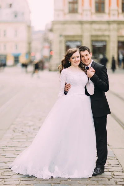 Pohledný ženich v černém obleku drží ruku nevěsty bílých šatech, staré slunné město pozadí — Stock fotografie