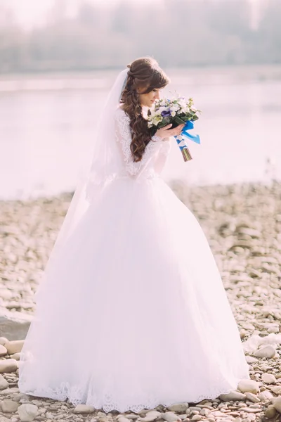 Portrait of young bride in white dress and veil sniffing wedding bouquet with blue bow outdoors — Stock Photo, Image