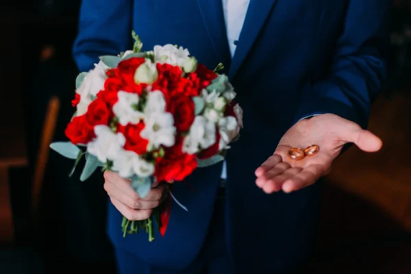 Traje azul vestido de hombre sosteniendo la novia y el novio anillos de boda con un ramo en otra mano — Foto de Stock