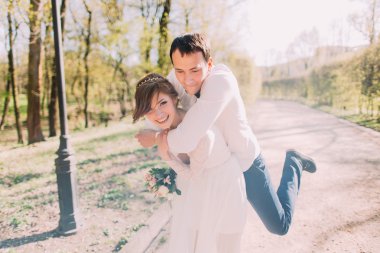 Smiling bride carrying on back groom outdoors in spring park clipart