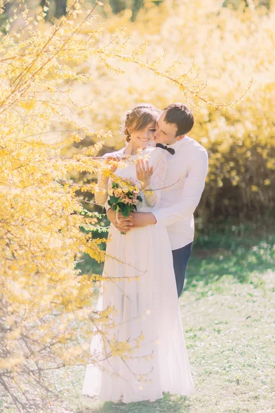 Jonge gelukkige bruidegom zachtjes omarmen achterkant van zijn mooie bruid in witte jurk buitenshuis met gele bloemen op achtergrond — Stockfoto