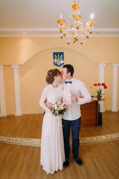Casal feliz de recém-casados se beijando no registro enquanto segurando a certidão de casamento — Fotografia de Stock