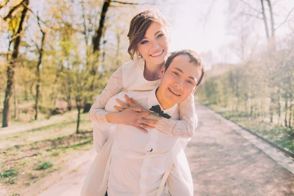 Zijn mooie blonde bruid draagt bruidegom glimlachend op rug buitenshuis in voorjaar park — Stockfoto
