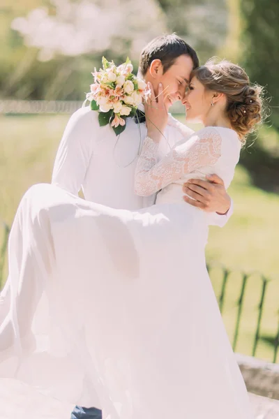 Sposo che porta la sua bella sposa sulle braccia appoggiate fronti al parco primaverile — Foto Stock