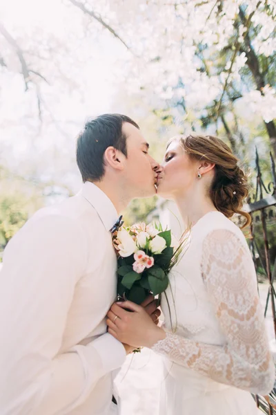 Giovane coppia di sposi che si baciano a vicenda tenendo bouquet di fiori di nozze in mano, all'aperto — Foto Stock