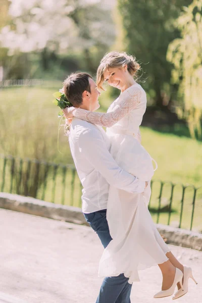 Handsome groom carrying beautiful laughing bride in his arms at spring park — Stock Photo, Image