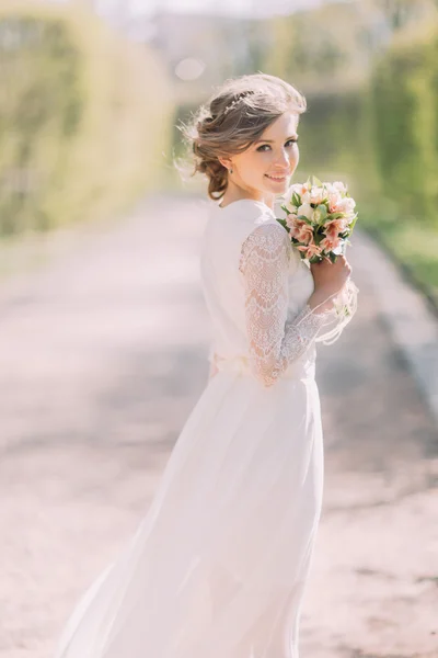 Bakifrån av unga blonda bruden i vit klänning med brudbukett stående utomhus — Stockfoto