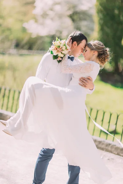 Groom carrying his beautiful bride on arms leaning foreheads at spring park — Stock Photo, Image