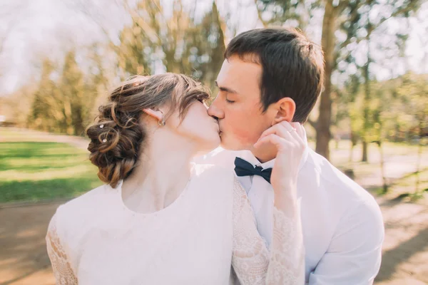 Beautiful happy young bride kissing handsome groom in sunlit park close-up