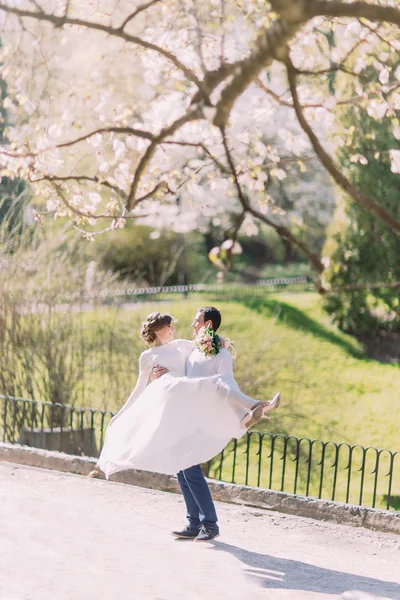 Beau marié portant belle mariée riante dans ses bras au parc de printemps — Photo