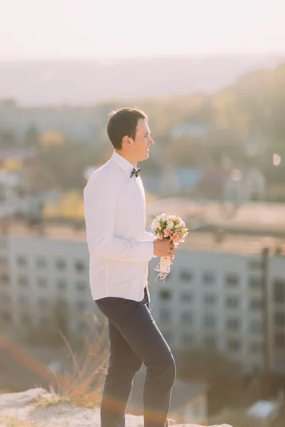 Bonito noivo em camisa branca e azul arco segurando buquê de casamento — Fotografia de Stock
