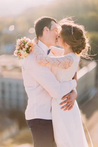 Счастливые молодожены целуются, пока невеста держит букет на фоне городского пейзажа — стоковое фото