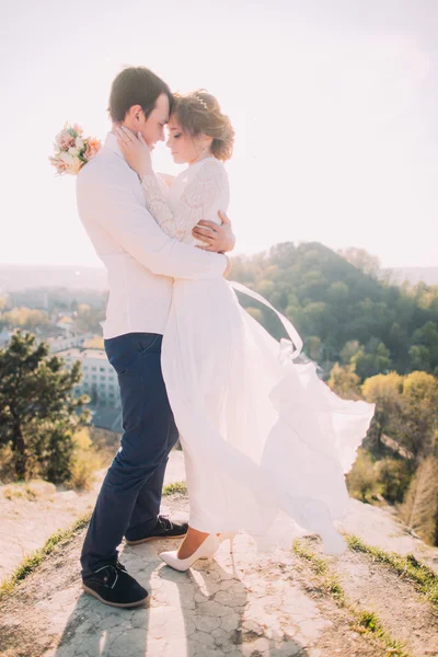 Atraente jovem casal amoroso de noivo e noiva gentil vestindo vestido branco fluttering no vento de pé no fundo ao ar livre ensolarado — Fotografia de Stock