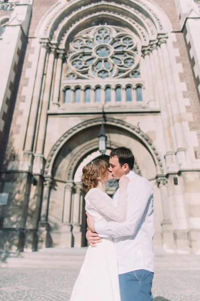 Feliz novia hermosa y elegante novio guapo besándose en el fondo de la iglesia edificio increíble en la ciudad — Foto de Stock