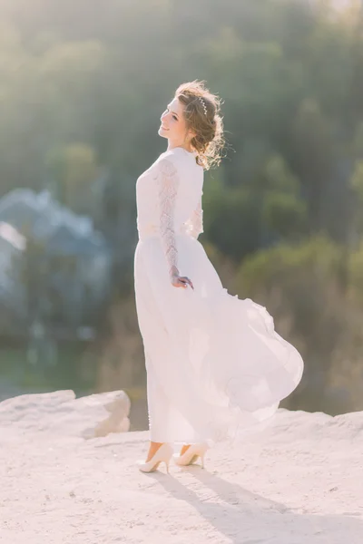 Bella sorridente giovane sposa bionda in piedi in cima alla collina sulla natura con abito bianco scorrevole — Foto Stock