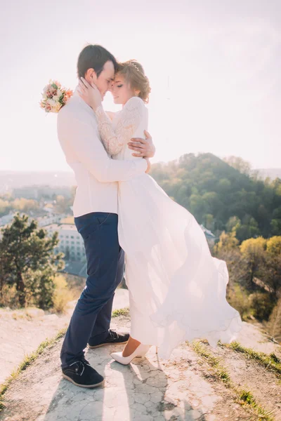 Attractive young loving couple of groom  and gentle bride wearing white dress fluttering in the wind standing on sunny outdoor background — Stock Photo, Image