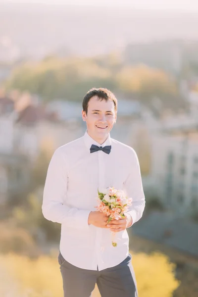 Elegante morena sonriente novio en camisa blanca con lazo azul sostiene lujoso ramo de flores al aire libre — Foto de Stock