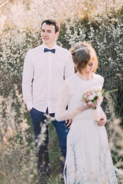 Красивый жених стоит позади невесты смотрит в сторону, его жена носит кружевное платье с букетом на переднем плане — стоковое фото