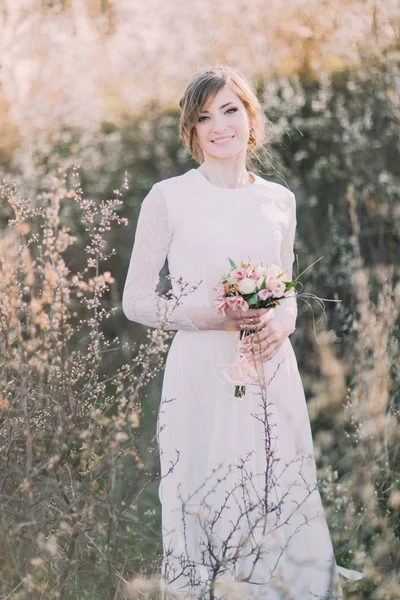 Νέοι όμορφη ξανθιά νύφη φορώντας λευκό φόρεμα με μπουκέτο σε καταπράσινο λιβάδι. Λεπτό κορίτσι απολαμβάνει τη φύση την άνοιξη — Φωτογραφία Αρχείου