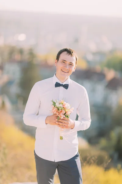Elegante morena sorrindo noivo em camisa branca com arco azul detém luxuoso buquê de flores ao ar livre — Fotografia de Stock