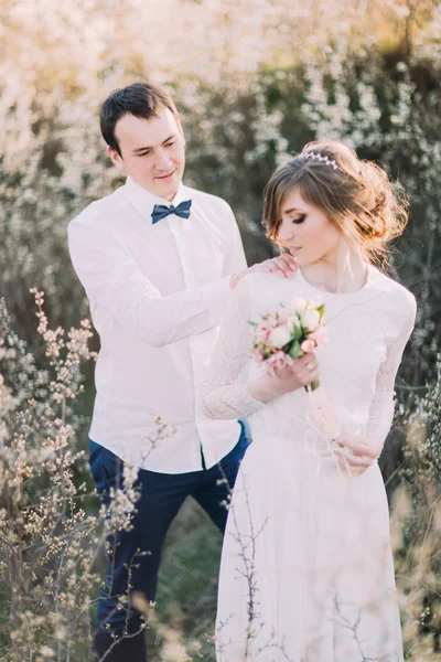 Knappe bruidegom staat achter bruid kijkt naar haar, raakt schouder van zijn vrouw dragen Lace Dress met boeket op de voorgrond — Stockfoto