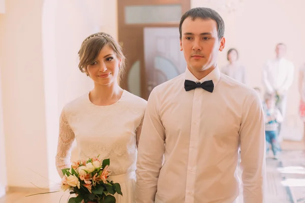 Χαμογελώντας ευτυχισμένη νύφη με Γαμήλια ανθοδέσμη και το γαμπρό στέκονται πιασμένοι χέρι χέρι στο ξενοδοχείο ή ληξιαρχείο γραφείο — Φωτογραφία Αρχείου
