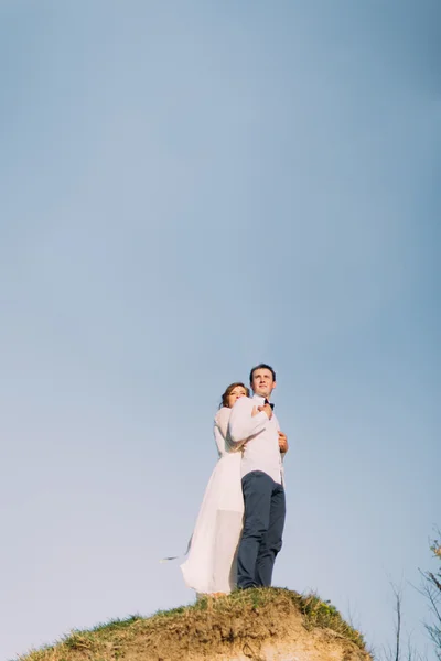 Glückliche schöne Braut umarmt ihren Bräutigam auf der Spitze des Hügels gegen den Himmel — Stockfoto