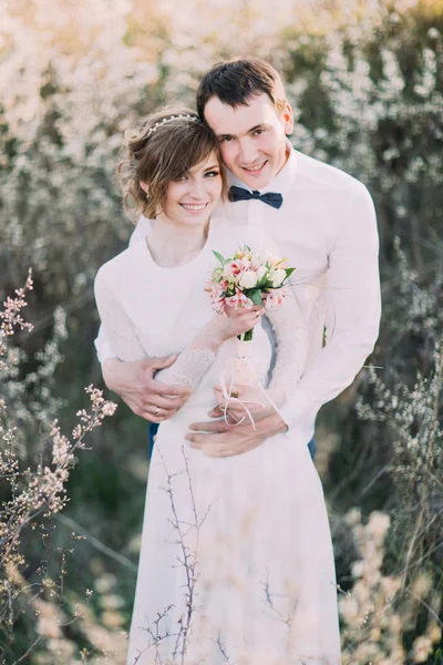 Schöner Bräutigam hält die Braut sanft mit Blumenstrauß im blühenden Frühlingsgarten zurück — Stockfoto