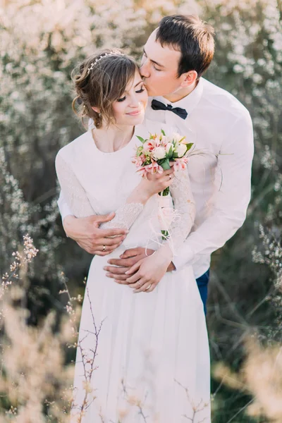 Schöner Bräutigam hält die Braut sanft zurück, hält Strauß und küsst Stirn im blühenden Frühlingsgarten — Stockfoto