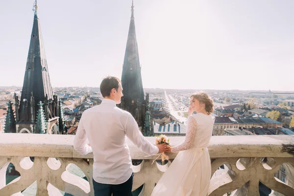 Bella sposa in abito bianco e bello sposo in piedi sul balcone con vista sulla città — Foto Stock