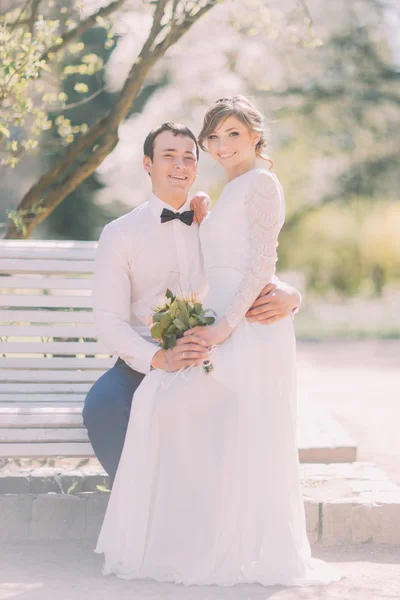 Cute young blonde bride sitting on grooms knees in spring park — Stock Photo, Image