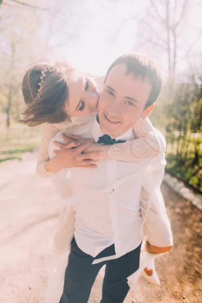 Novio sonriente lleva a su hermosa novia rubia en la espalda al aire libre en el parque de primavera —  Fotos de Stock