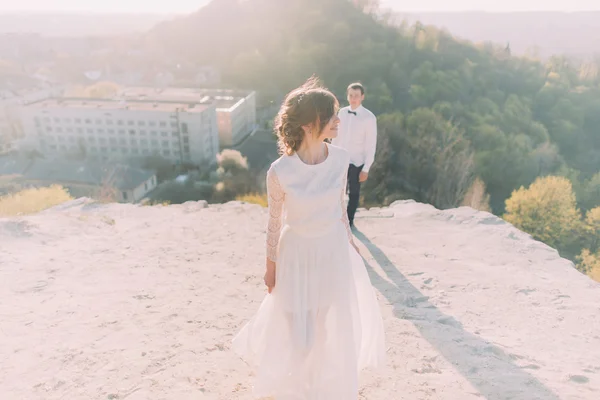 Junger Bräutigam in weißem Hemd und Schleife steht hinter der tanzenden schönen Braut im Brautkleid — Stockfoto