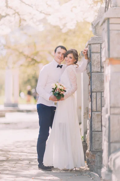 Jeune mariée et marié étreignant avec bouquet de fleurs de printemps près de la clôture — Photo