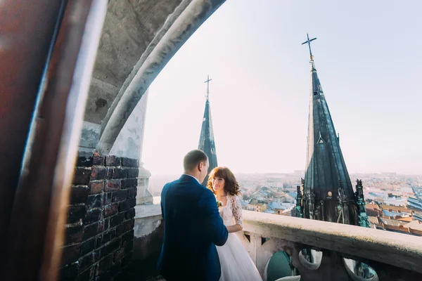 Нежная красивая невеста и жених держатся за руки, обнимаясь лицом к лицу на древнем балконе, фоне городского пейзажа — стоковое фото