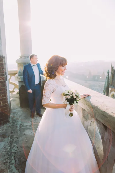 Beautiful bride wearing white dress posing on old balcony, groom in background — Stock Photo, Image