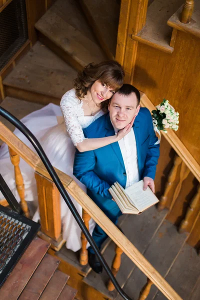 Прекрасная чувственная невеста обнимает из-за своего красивого жениха, держа книгу на деревянной лестнице старой библиотеки. — стоковое фото