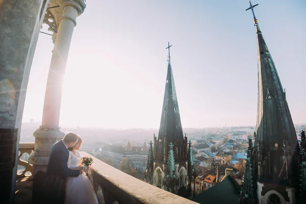 Gyönyörű pár, a menyasszony és a vőlegény pózol a régi erkély oszloppal, városkép háttér — Stock Fotó