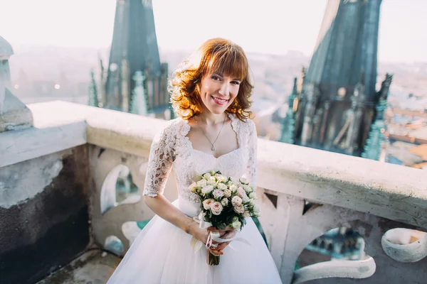 Невеста смотрит в камеру, держа букет на балконе с большим городским пейзажем — стоковое фото