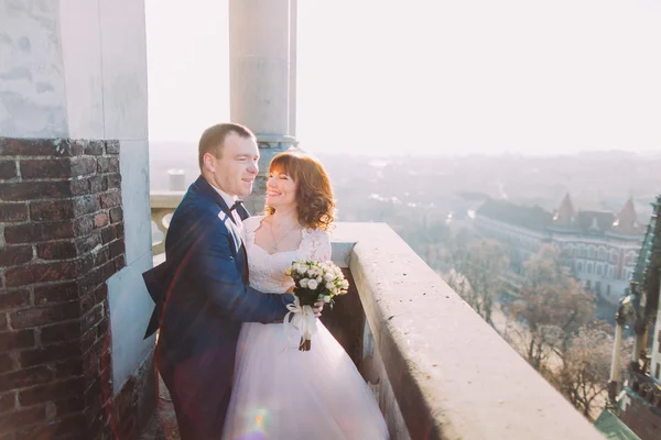都市の背景を持つ古い城のバルコニーで新婚の花嫁を後ろから抱き締めるハンサムな官能的な新郎 — ストック写真